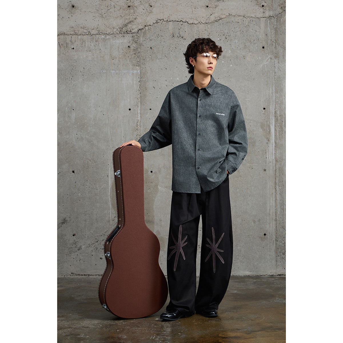 Model wearing a charcoal button-up shirt with small white chest lettering and black wide-leg trousers with starburst designs.