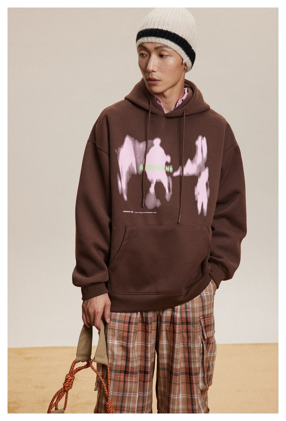 Model wearing a brown graphic hoodie with a beanie and plaid cargo pants, holding a rope bag
