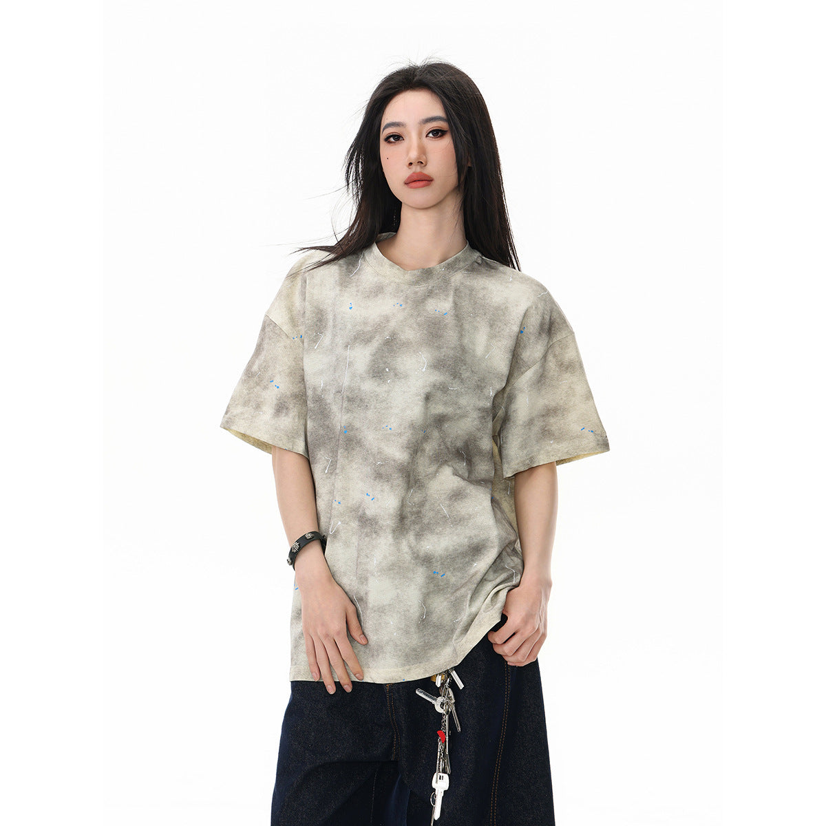 Model wearing an oversized beige-gray mottled T-shirt with small scattered flecks and dark jeans.