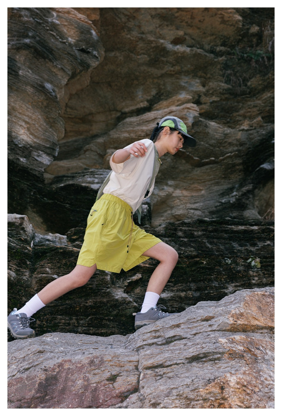 Woman wearing a white T-shirt, lime-green shorts and cap while striding across a rocky trail.