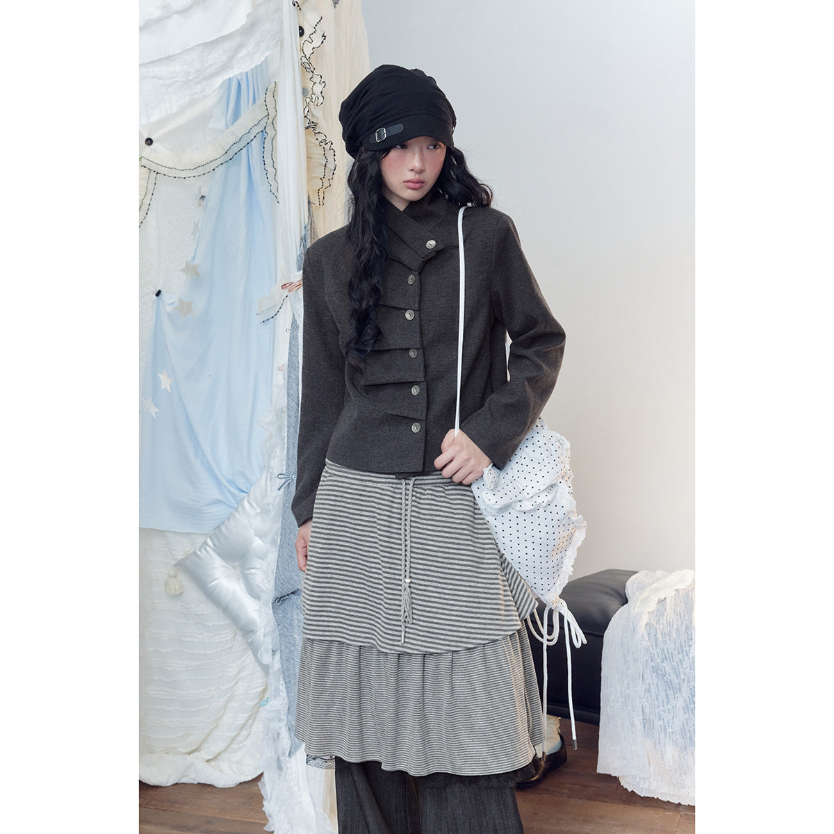 Model wearing a charcoal asymmetrical-button jacket, layered gray striped skirt and dark wide-leg pants with a black cap.