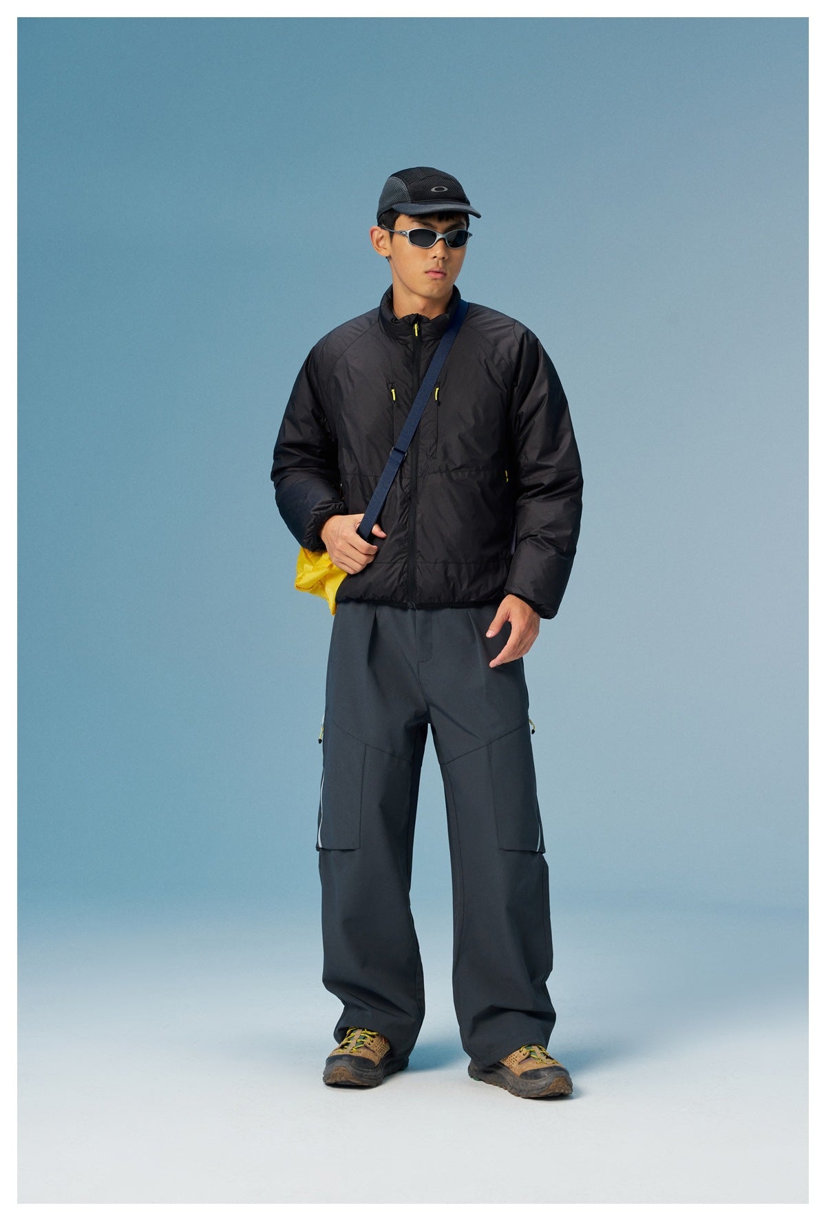 Model wearing the black zip jacket with gray cargo pants, a cap, sunglasses and a yellow crossbody bag, front view