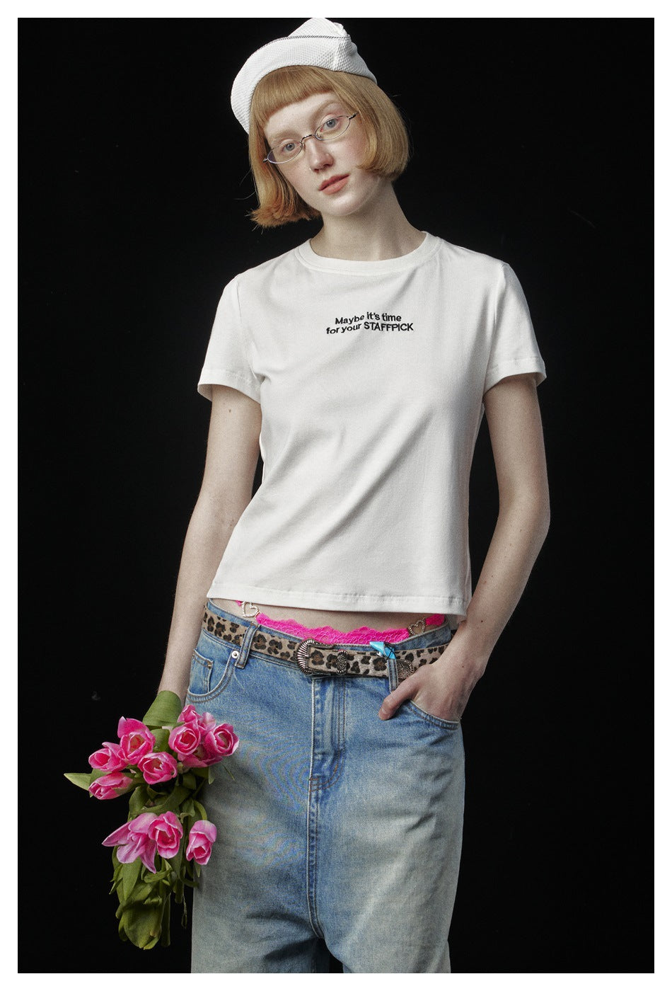 Model in a white slogan T-shirt and blue jeans, shown from the waist up with pink tulips against black.
