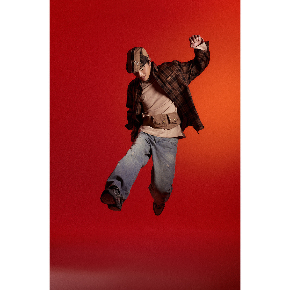Model in a dark plaid button-up shirt, taupe top, and loose blue jeans, posing against a red backdrop.