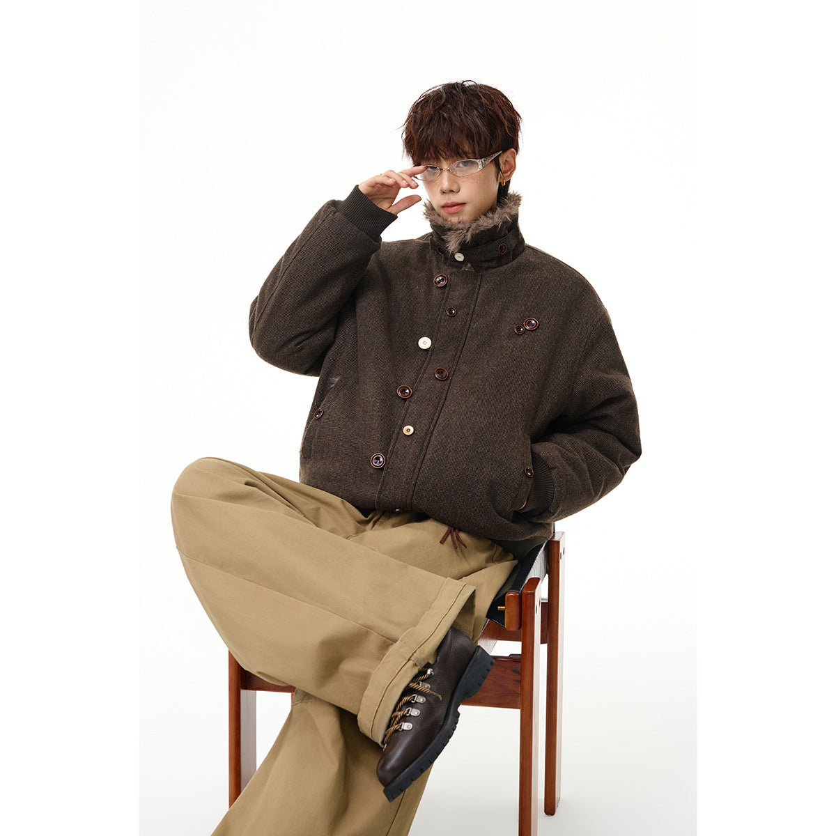 Model seated in a dark brown jacket with a high fuzzy collar, contrast buttons and ribbed cuffs, paired with tan trousers.