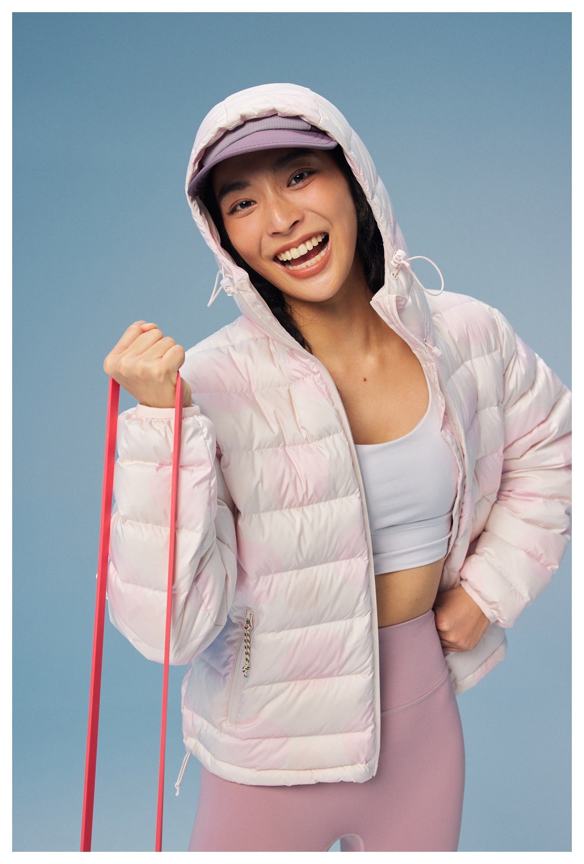 Smiling model wearing an open pale pink hooded puffer jacket with a subtle circular pattern and lavender leggings.