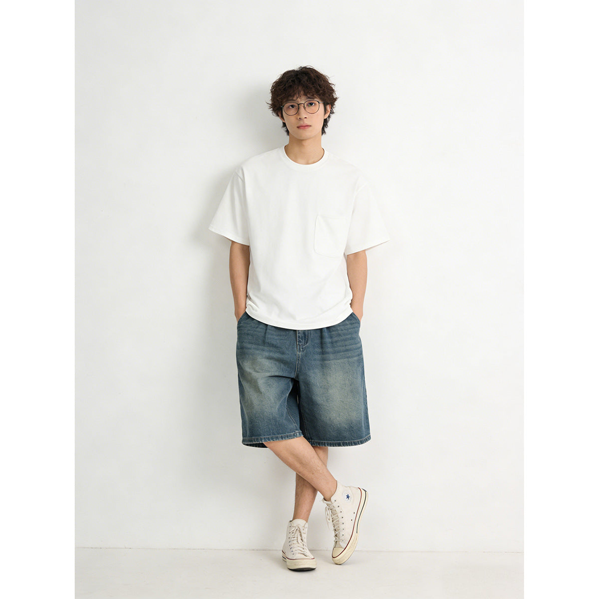 Model wearing a loose white crew-neck T-shirt with a chest pocket and blue denim shorts.