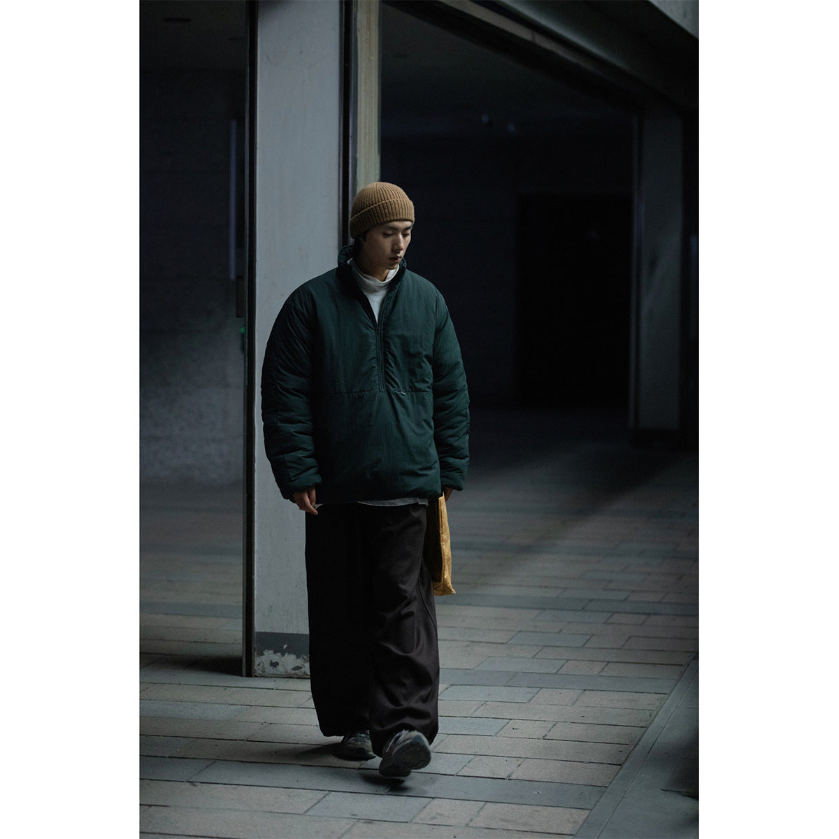 Full-length view of a model in a dark teal padded pullover jacket, tan beanie and loose black trousers.