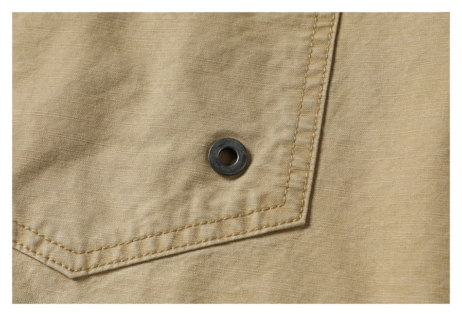 Extreme close-up of the metal eyelet and double stitching on the tan shirt's chest pocket.