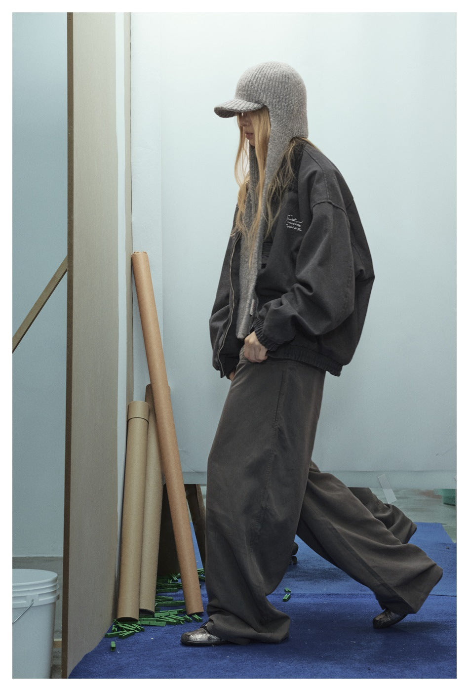 Side view of a model in an oversized charcoal zip jacket and wide-leg pants, with a gray knit cap and long scarf.