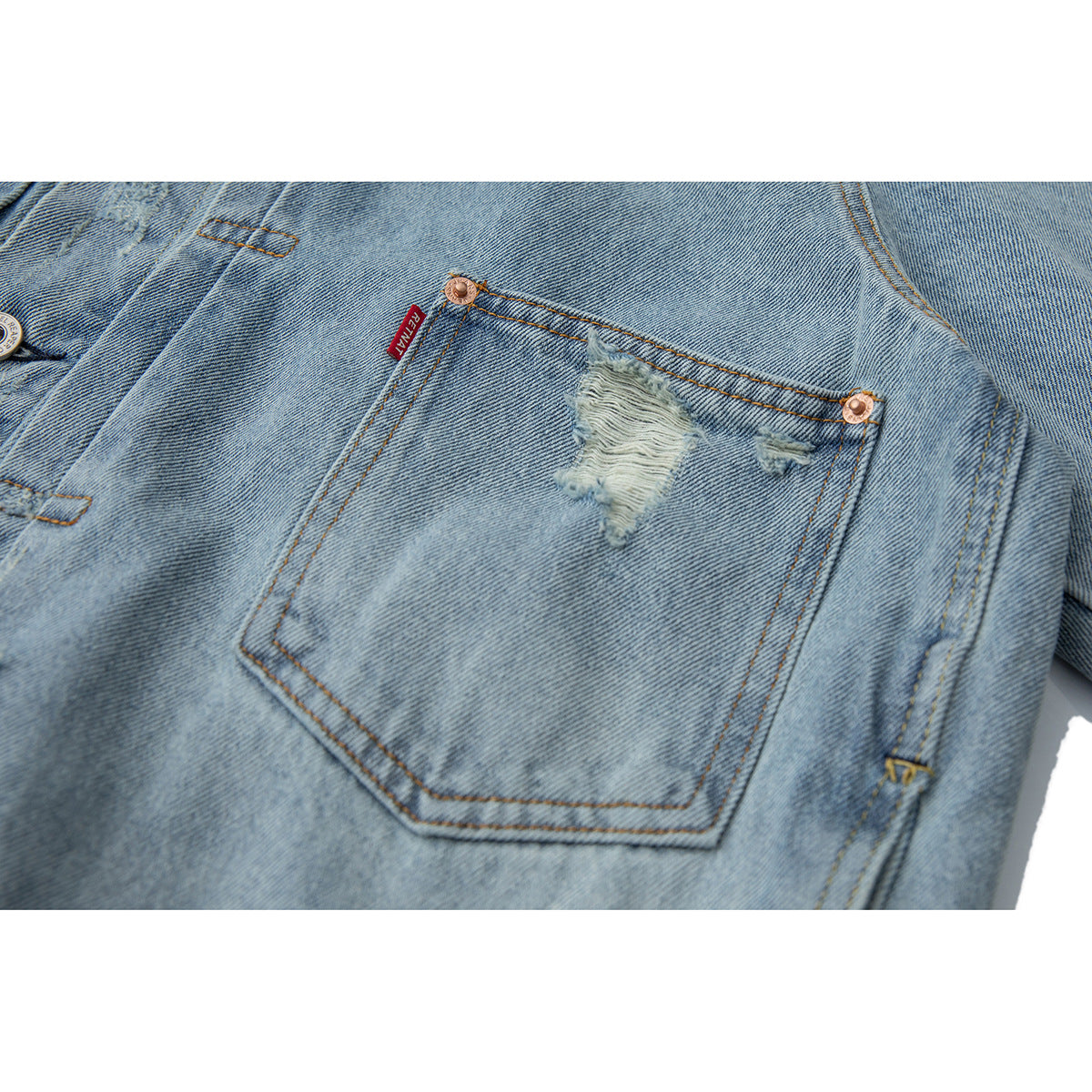 Close-up of a distressed patch pocket on light blue denim, with copper rivets and a red fabric tab.