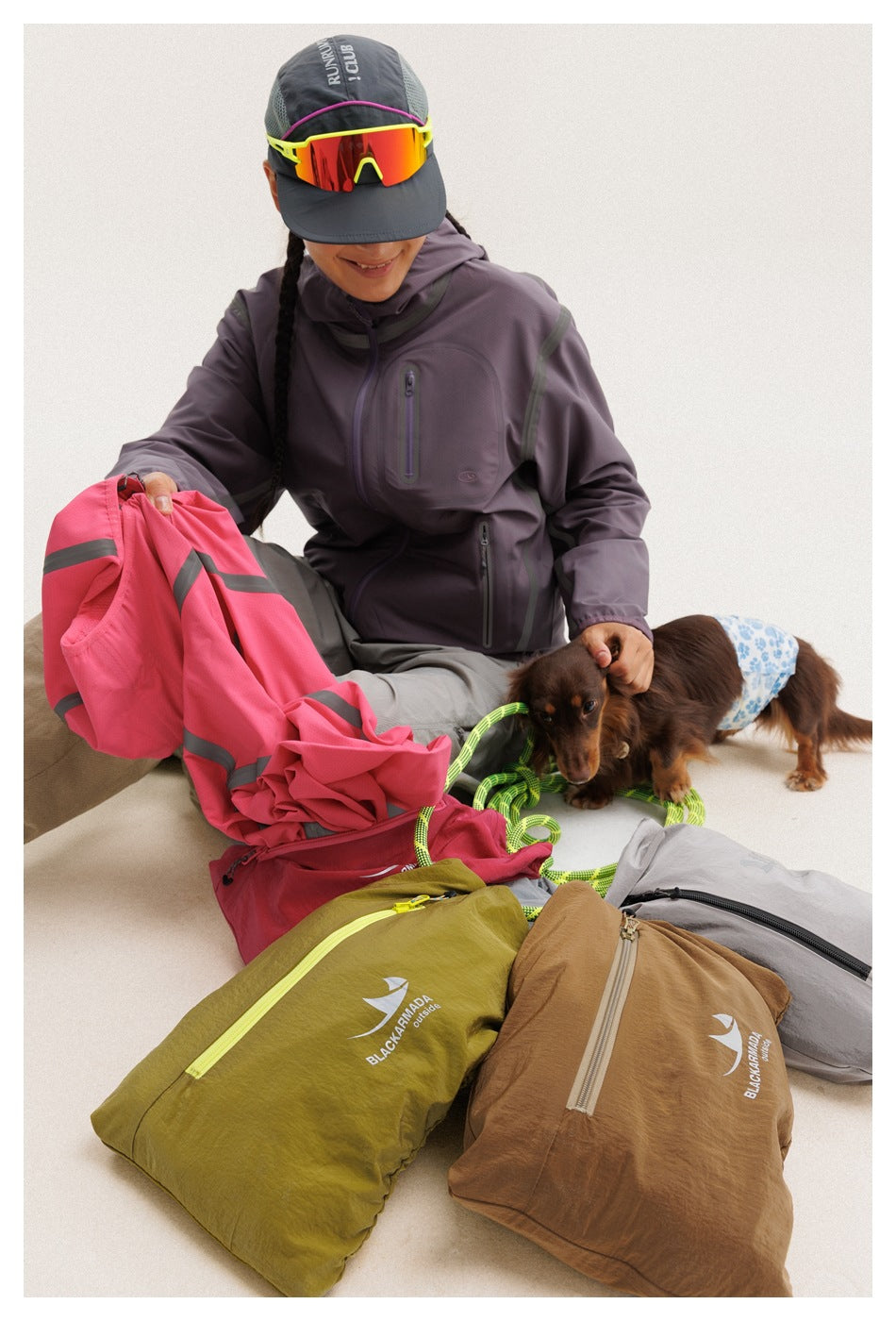 Seated model in a dark plum jacket with a pink jacket and assorted bags arranged around them.