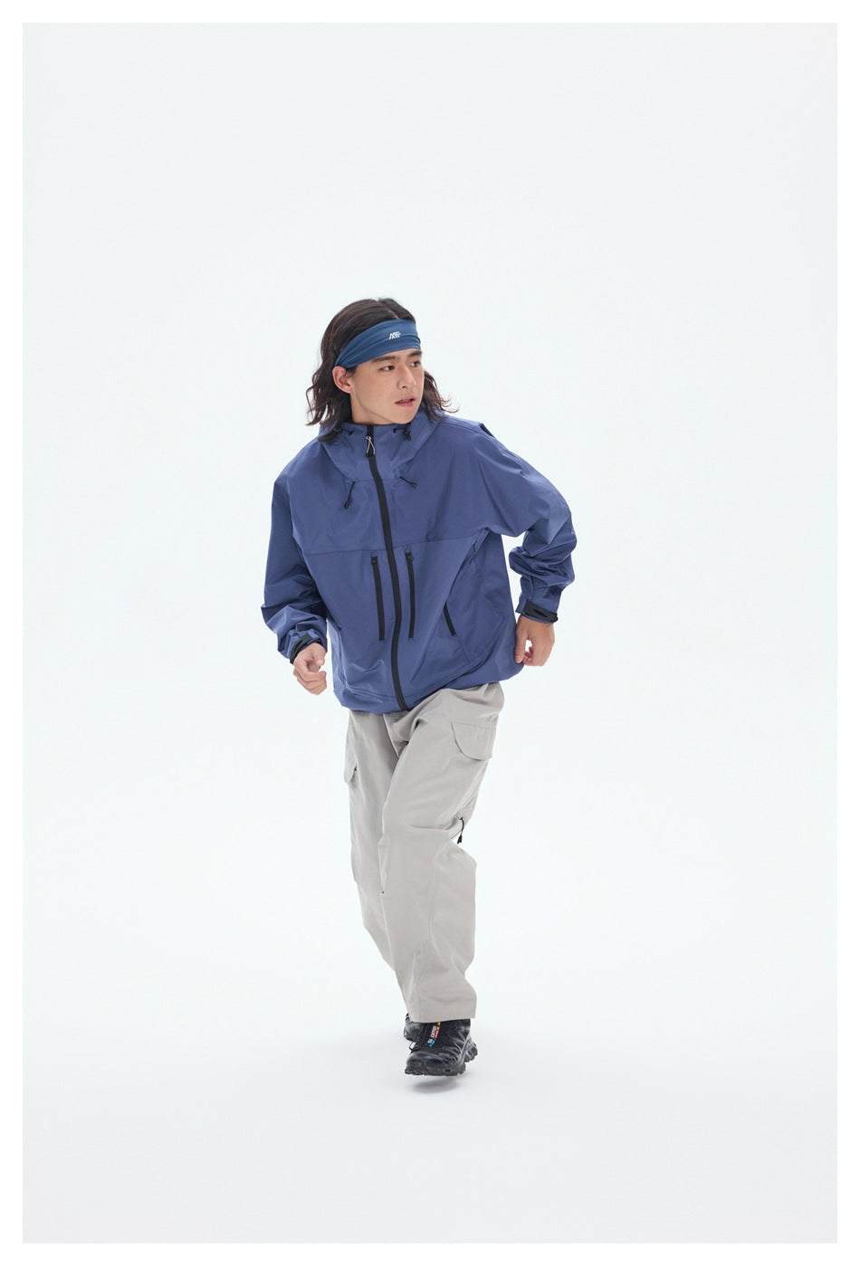 Model wearing a blue hooded jacket with black zippers and light gray cargo pants in a walking pose.