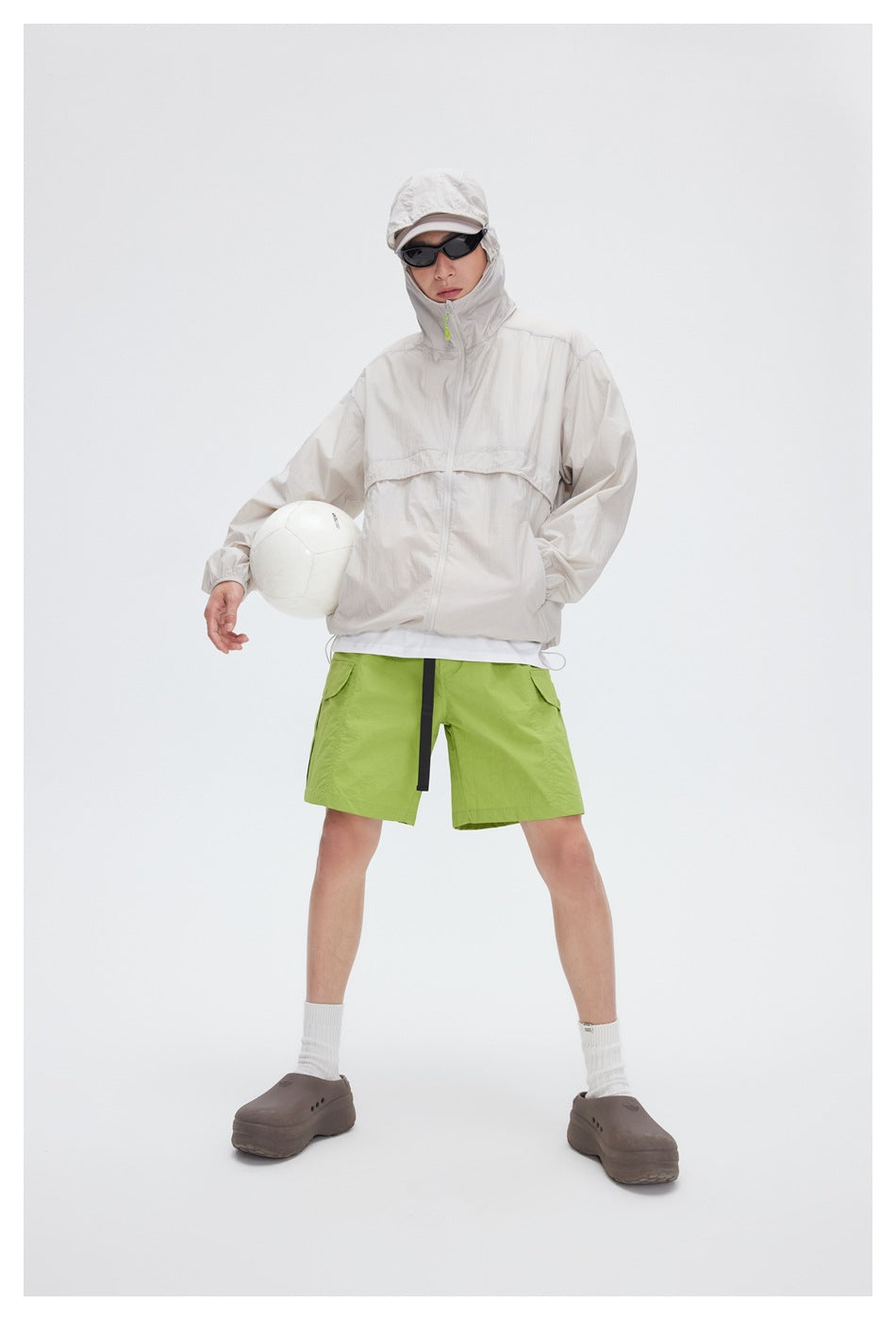 Front view of a model in a pale beige hooded jacket and lime-green shorts, holding a white ball.