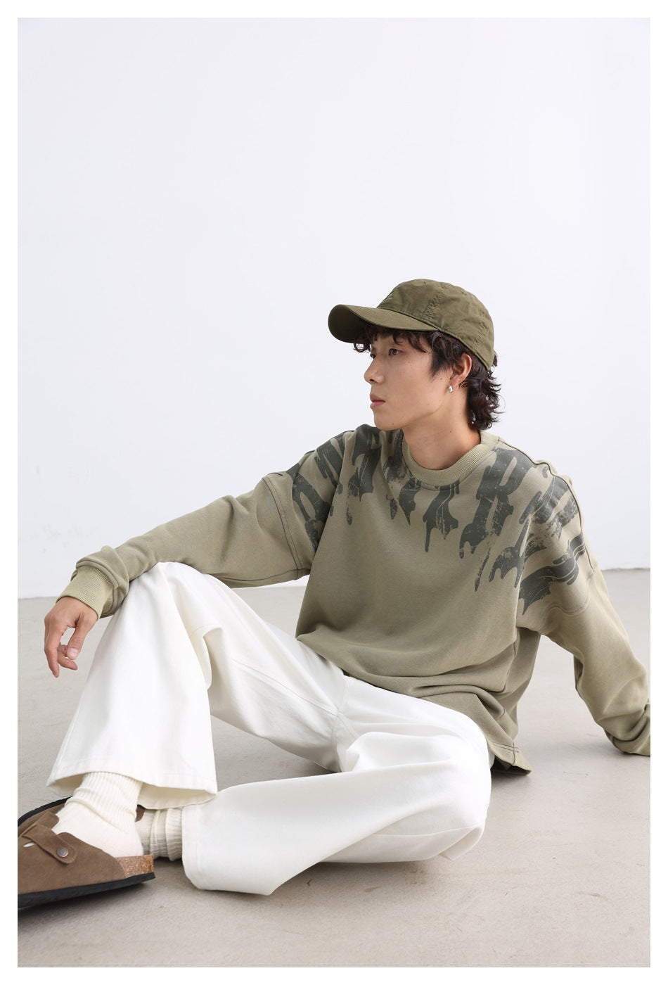 Person seated on the floor wearing the green printed sweatshirt with a cap, white pants and brown clogs, side view