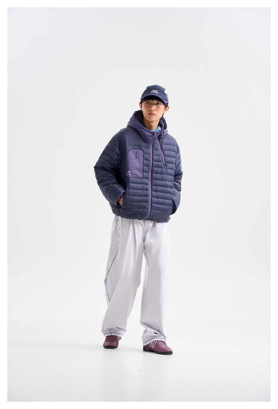 Full-length view of a person wearing a navy quilted jacket with purple hood and pocket, white trousers and purple shoes.