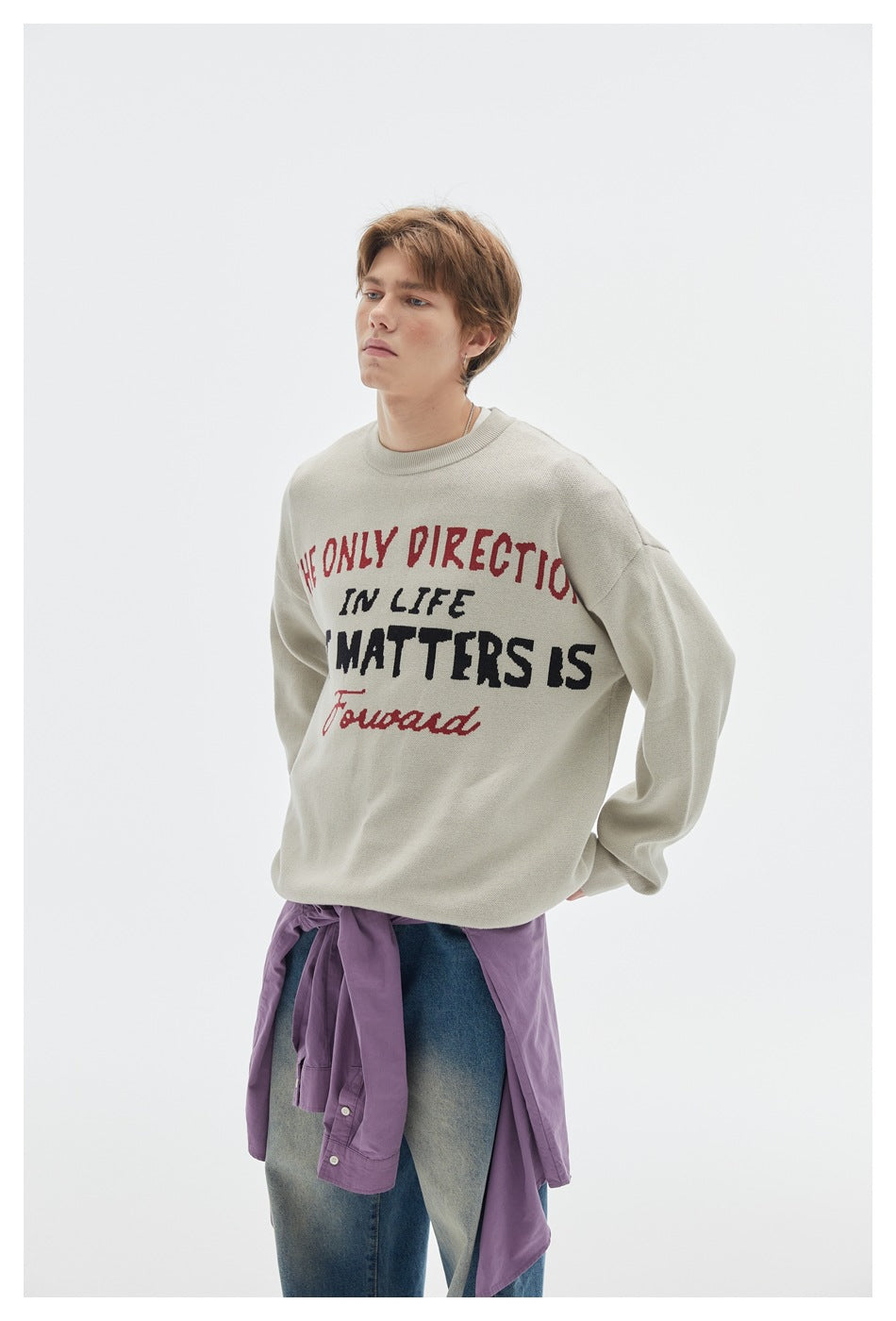 Model wearing a light gray crewneck sweater with red and black lettering, blue jeans, and a purple shirt tied at the waist.