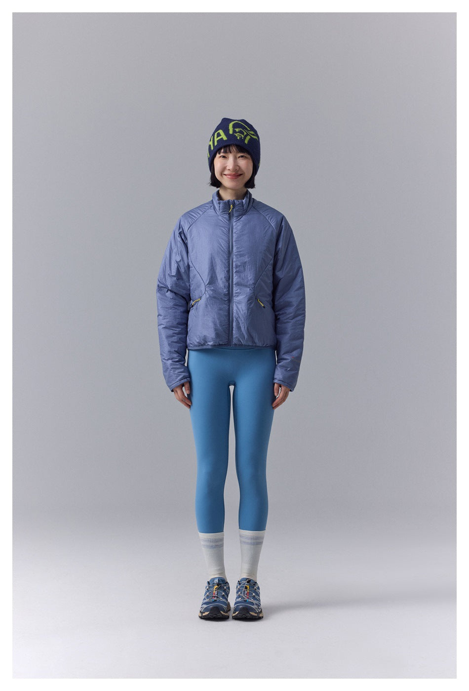 Model wears a blue-gray zip-front jacket with blue leggings and a navy beanie.