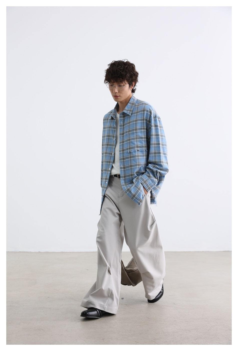 Model walking in a loose blue plaid shirt with a frayed chest pocket and hem, paired with light wide-leg trousers.