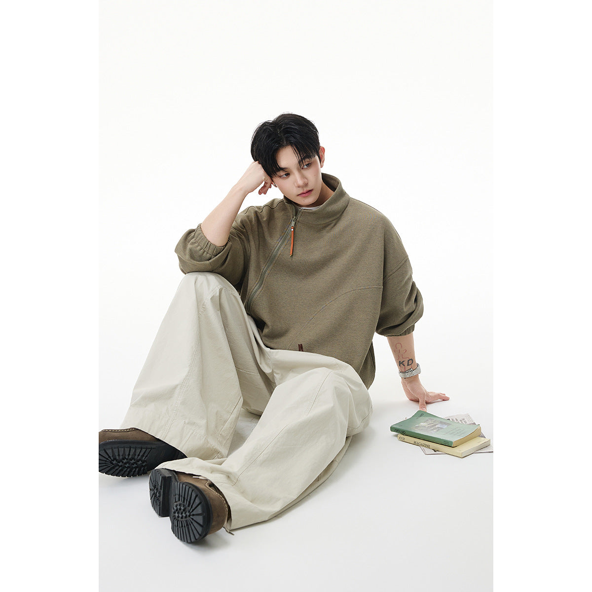 Model seated in an olive-gray oversized zip pullover with a tall collar and cream wide-leg trousers.
