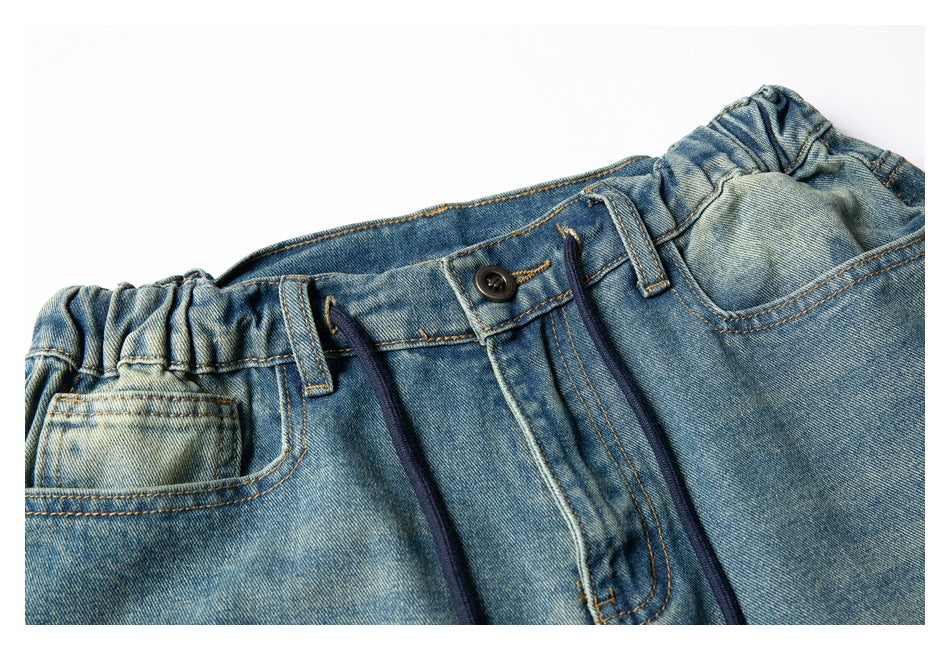 Angled close-up of blue denim trousers showing the elastic waistband, front button, drawcords and pocket.