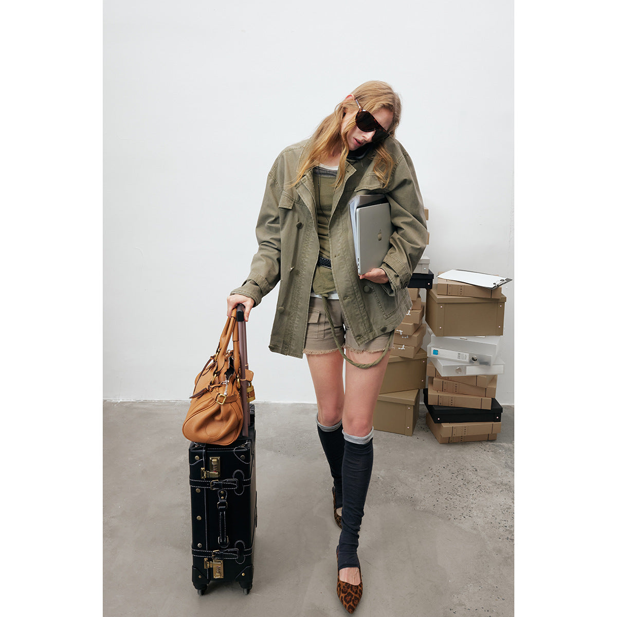 Model wearing an oversized olive utility jacket with flap pockets and buttons, paired with shorts and dark leg warmers.