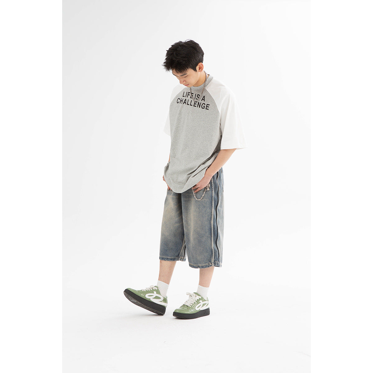 Model in a gray raglan T-shirt with white sleeves and black lettering, loose washed denim shorts, and green sneakers.