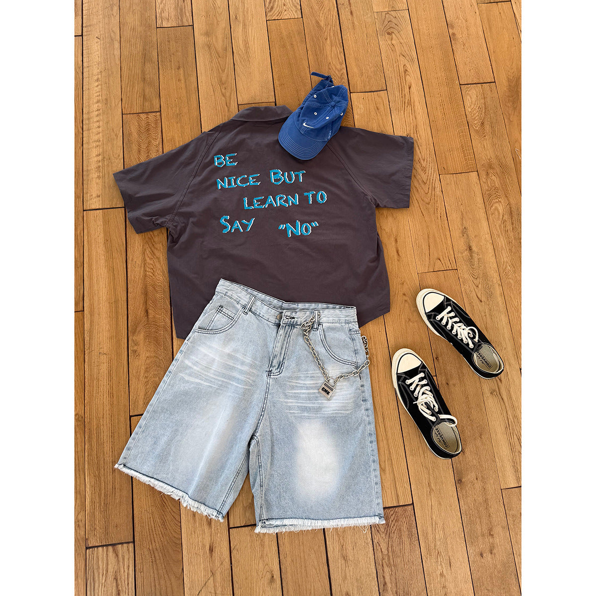 Flat lay of a charcoal short-sleeve shirt with blue lettering on the back, light denim shorts, a blue cap and black sneakers.