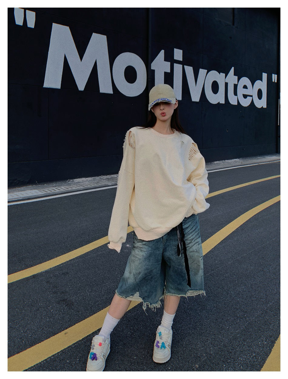 Model wearing an oversized cream distressed sweatshirt and frayed denim shorts with a cap and sneakers on a street.