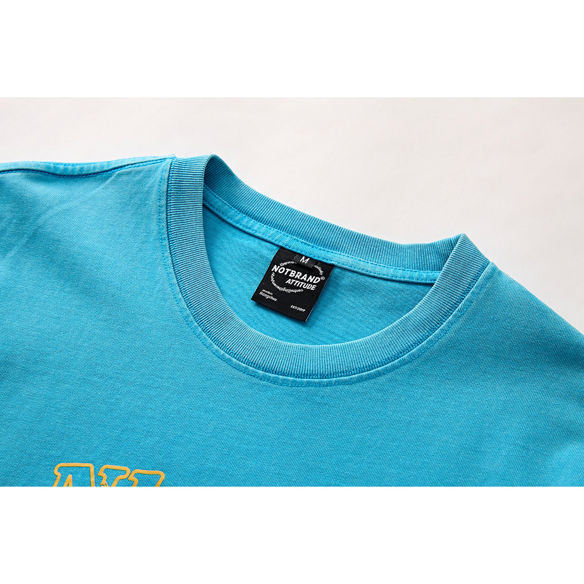 Close-up of a turquoise T-shirt neckline with a black neck label and part of a yellow chest graphic.