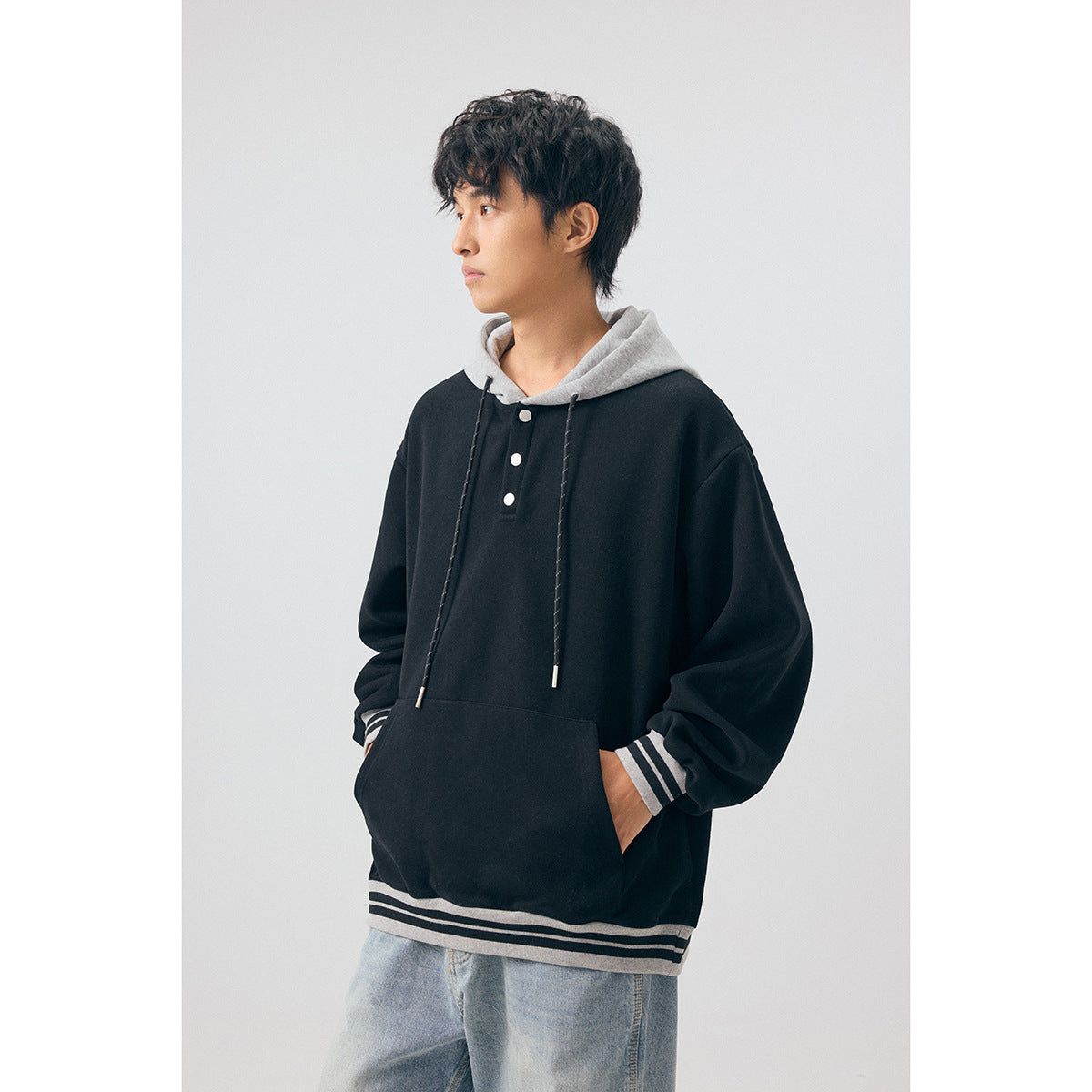 Model wearing a black pullover hoodie with a gray hood, three-button placket, front pouch pocket, and striped cuffs and hem.