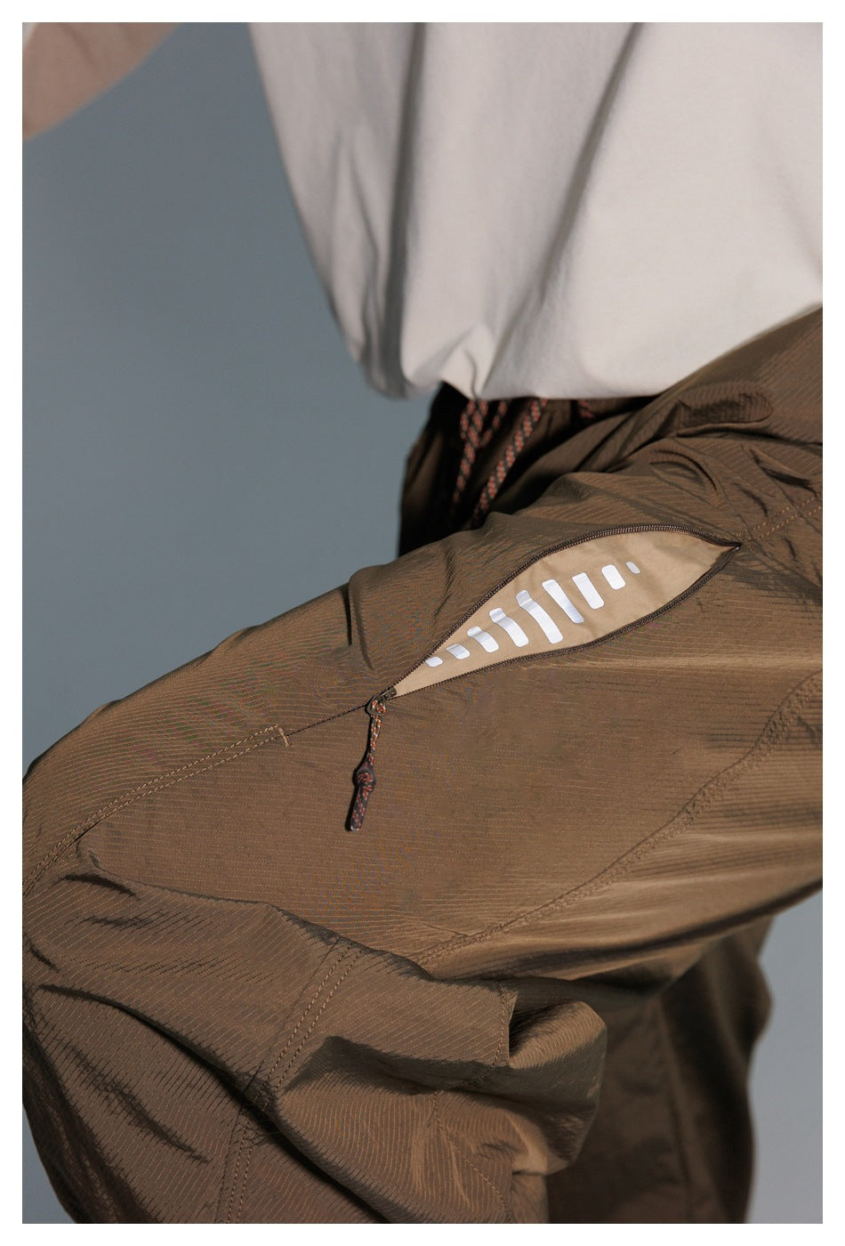 Close-up of olive-brown pants showing a zippered pocket with a light tan panel and white bar-shaped design.