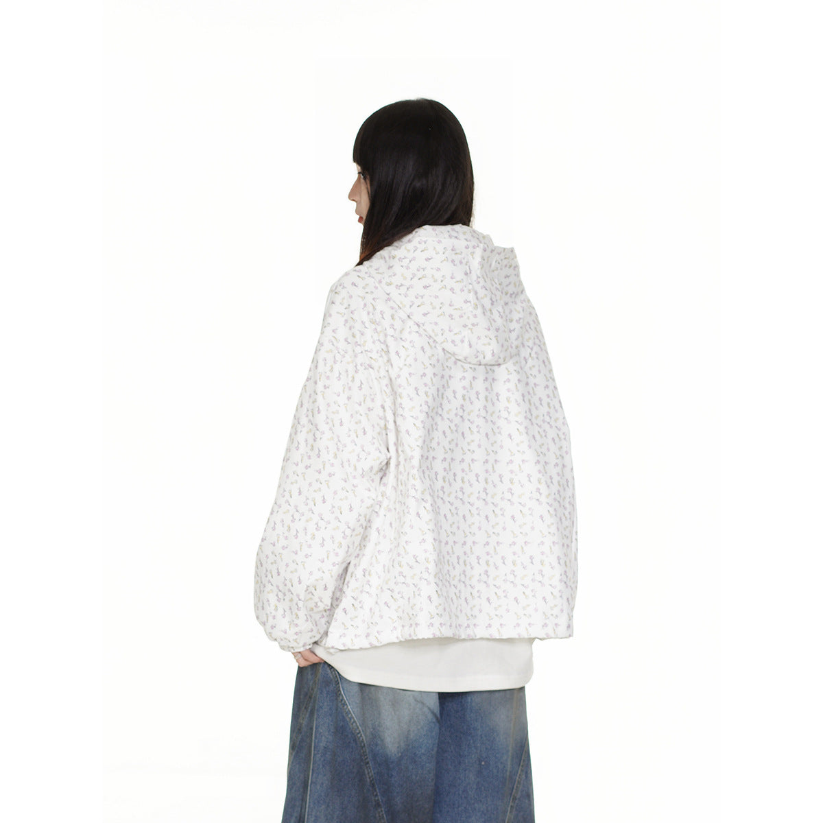 Rear view of a model wearing a white jacket with a tiny floral print and a hood, over loose blue jeans.