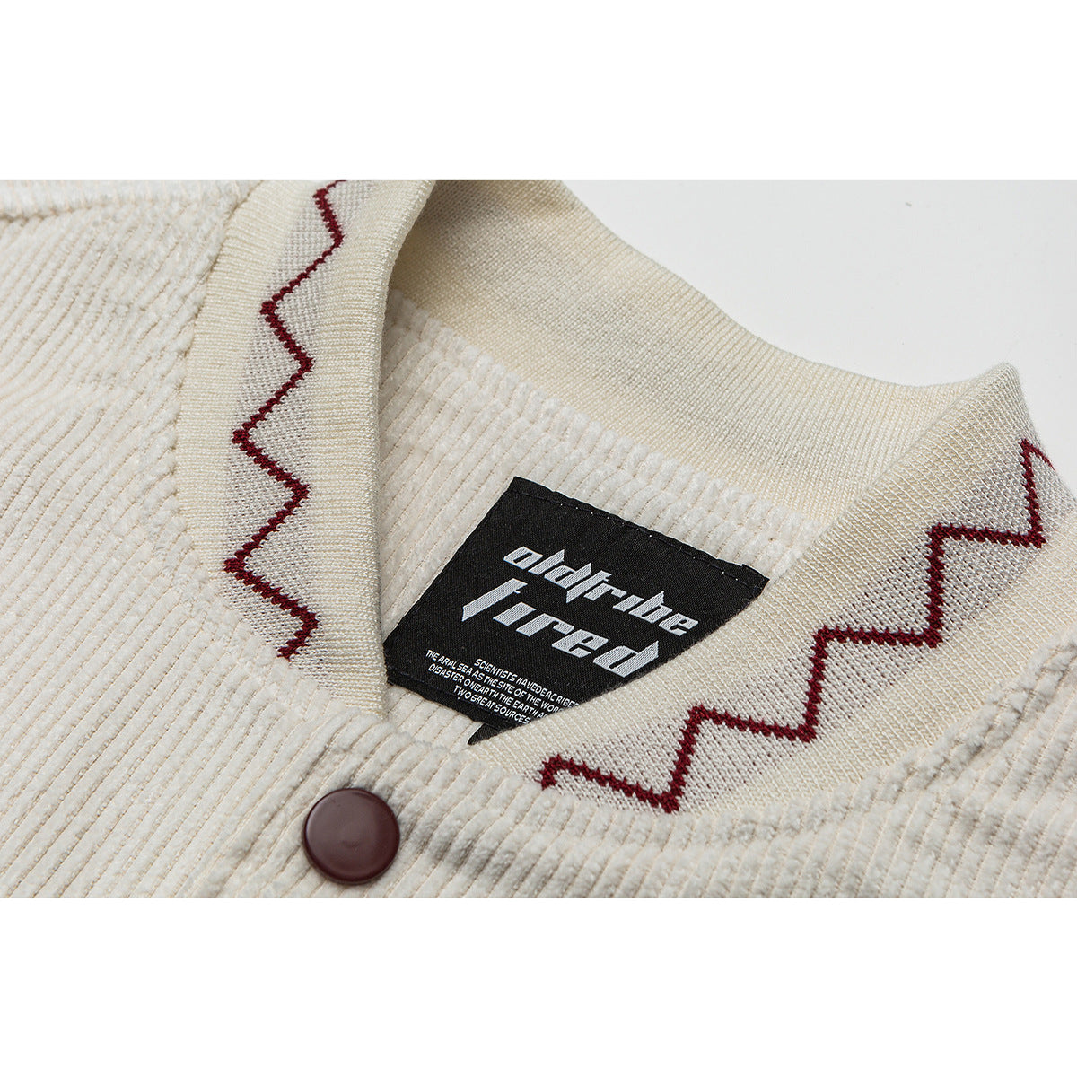 Close-up of a cream ribbed jacket collar with burgundy zigzag trim, a brown button, and a black neck label.
