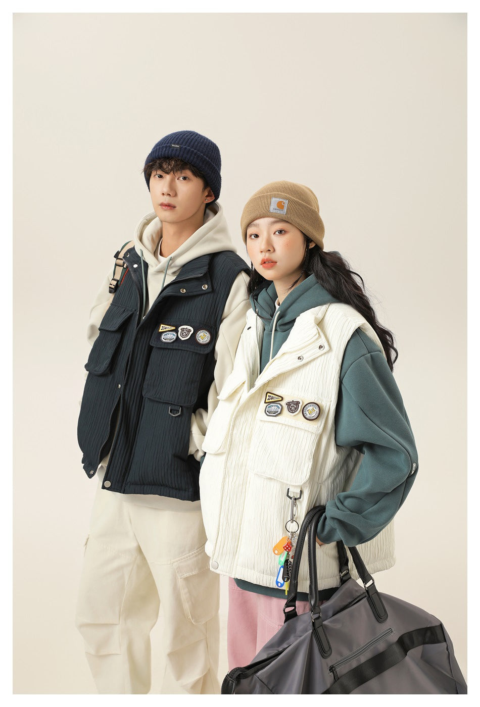 Two models pose in navy and cream utility vests over hoodies; the cream vest has large flap pockets and patches.