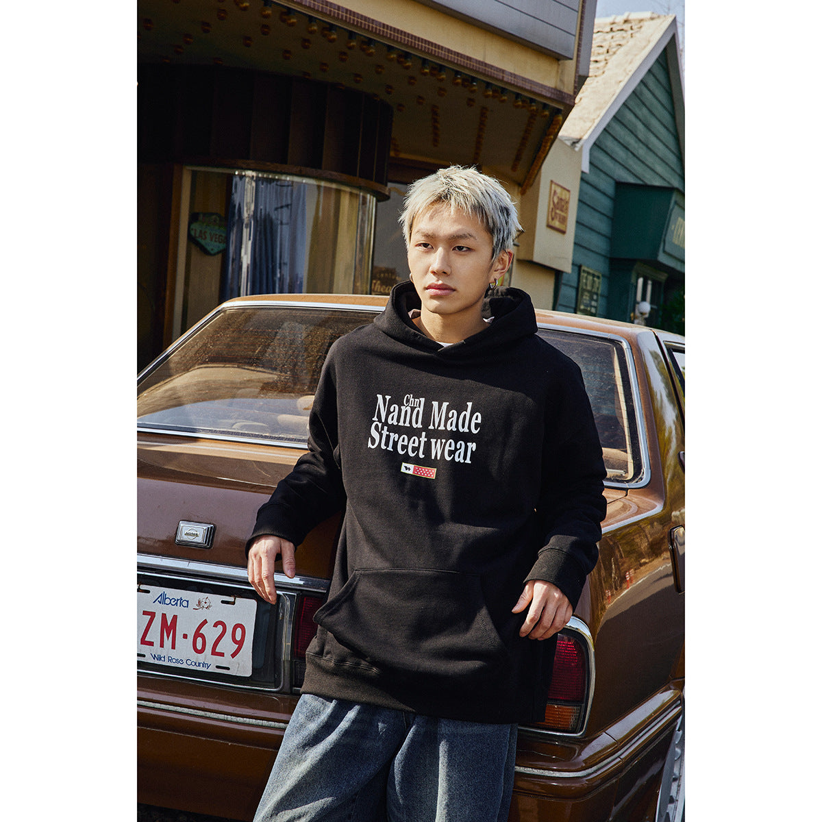 Model wearing a black graphic pullover hoodie with a kangaroo pocket and drawstring hood, posed beside a car.