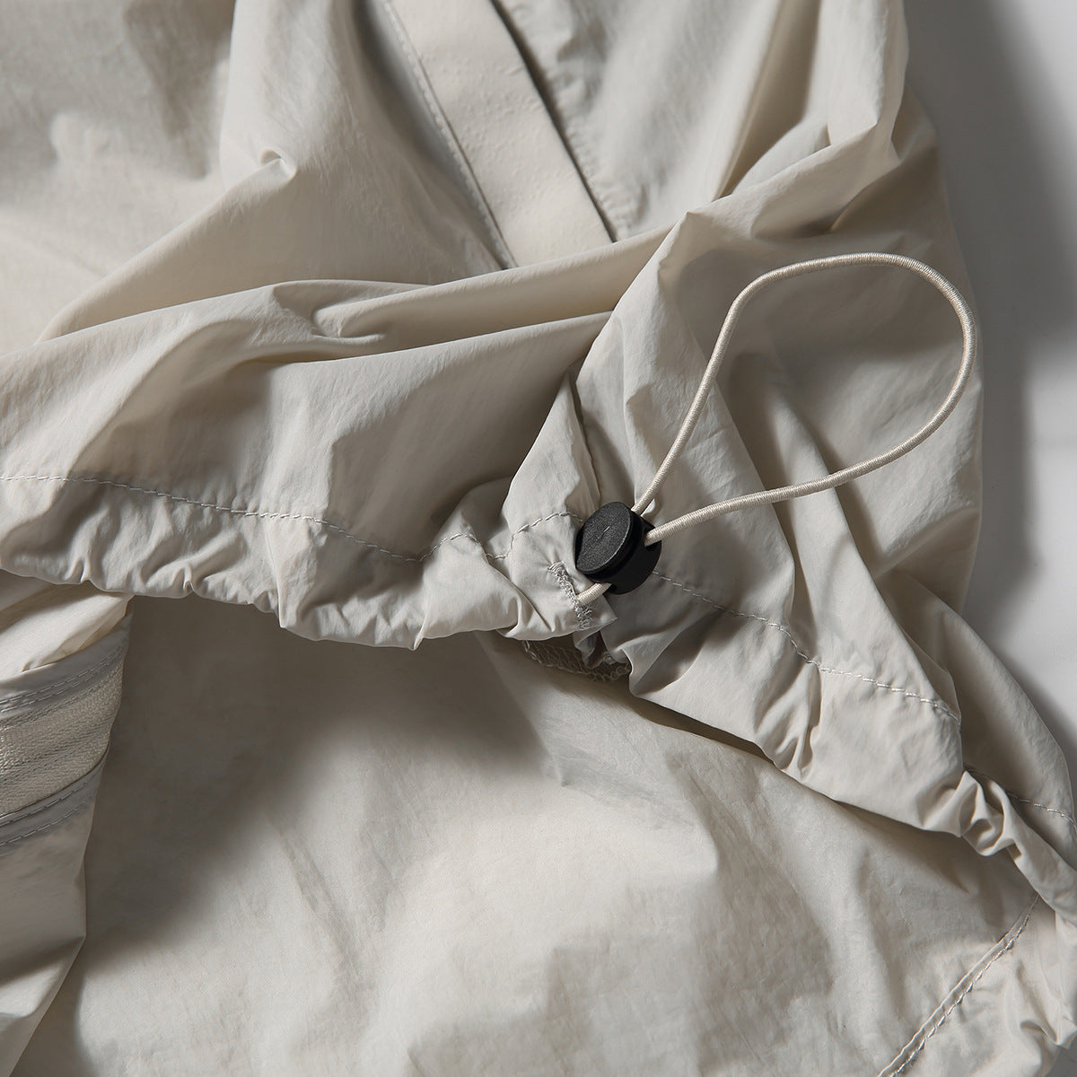 Close-up of the light beige jacket's hem, showing a black cord toggle and elastic drawcord.