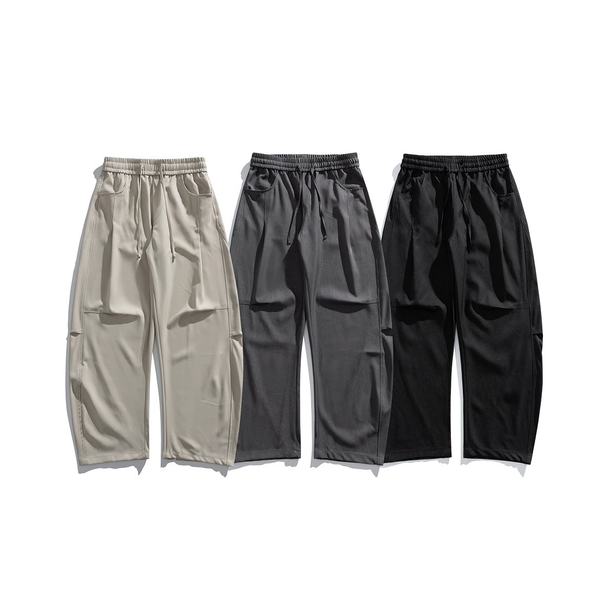 Three pairs of loose straight-leg trousers with elastic waistbands and drawstrings in beige, charcoal gray, and black.