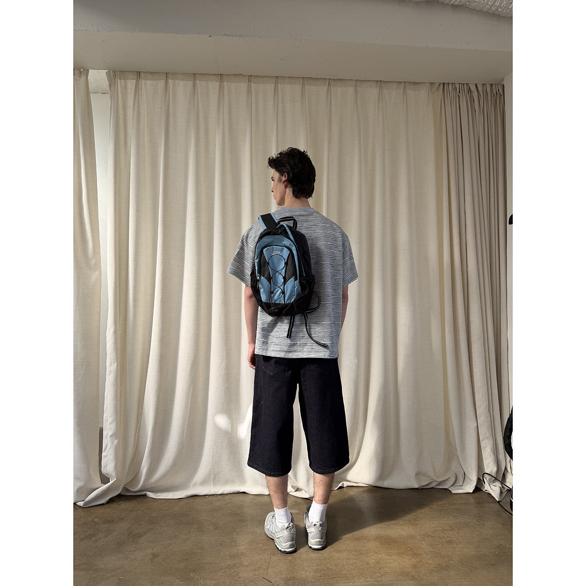 Rear view of a model wearing a gray striped T-shirt and black knee-length shorts with a blue-and-black backpack.