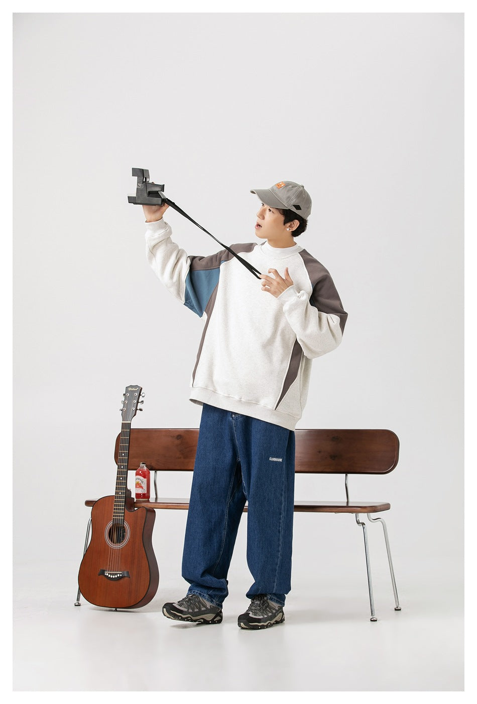 Model in light gray colorblock sweatshirt with brown and blue panels, jeans and cap, standing beside a guitar and bench