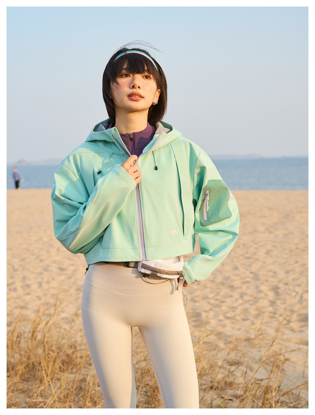 Beachside view of a model wearing a mint hooded cropped jacket with lavender zipper trim and cream leggings.