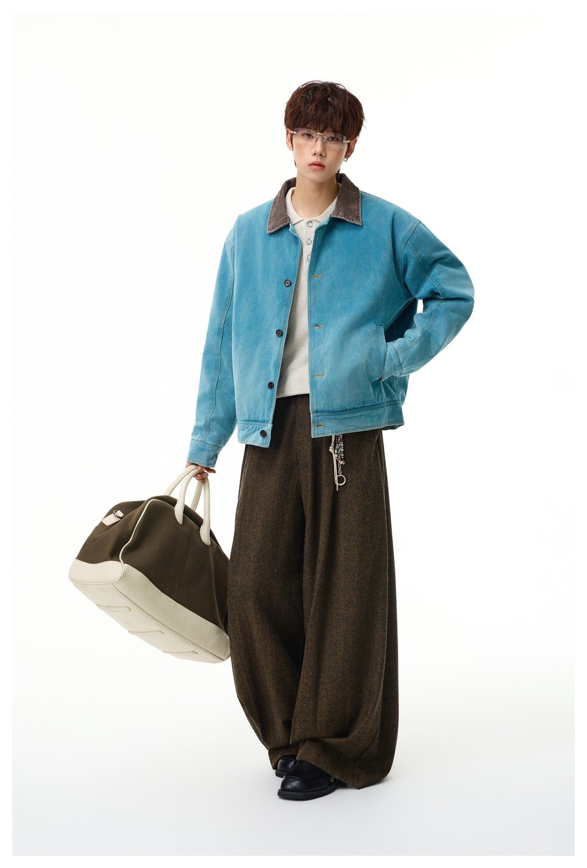 Model wearing the teal-blue washed jacket with a cream knit polo, brown wide-leg trousers and a large duffle bag