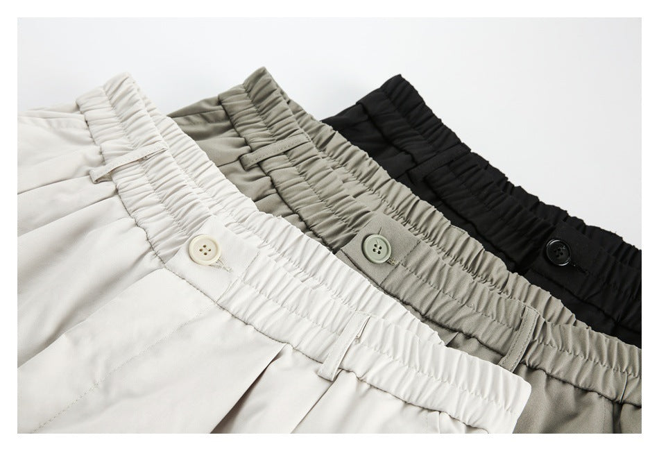 Three pairs of trousers in off-white, gray-khaki, and black, shown with gathered elastic waistbands and buttons.