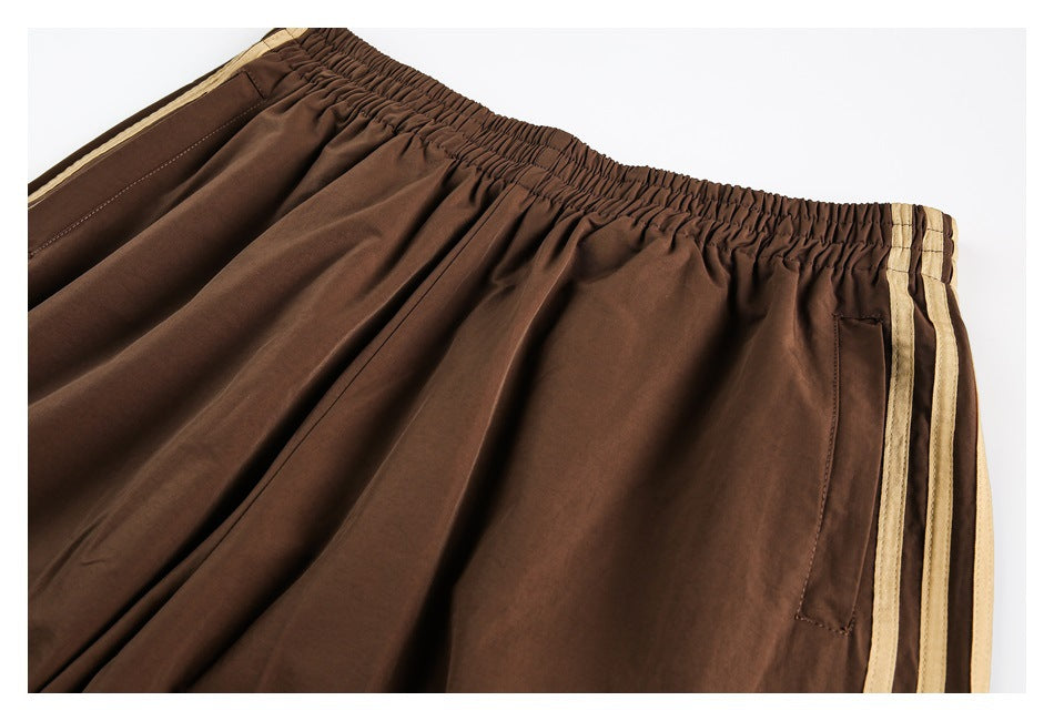 Close-up of brown track pants showing the gathered elastic waistband, side pocket, and three tan stripes.