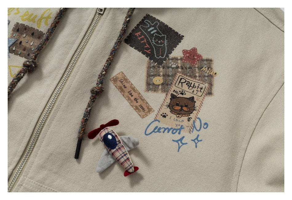 Close-up of playful printed patches and a small plaid airplane charm on a beige zip-front hoodie.