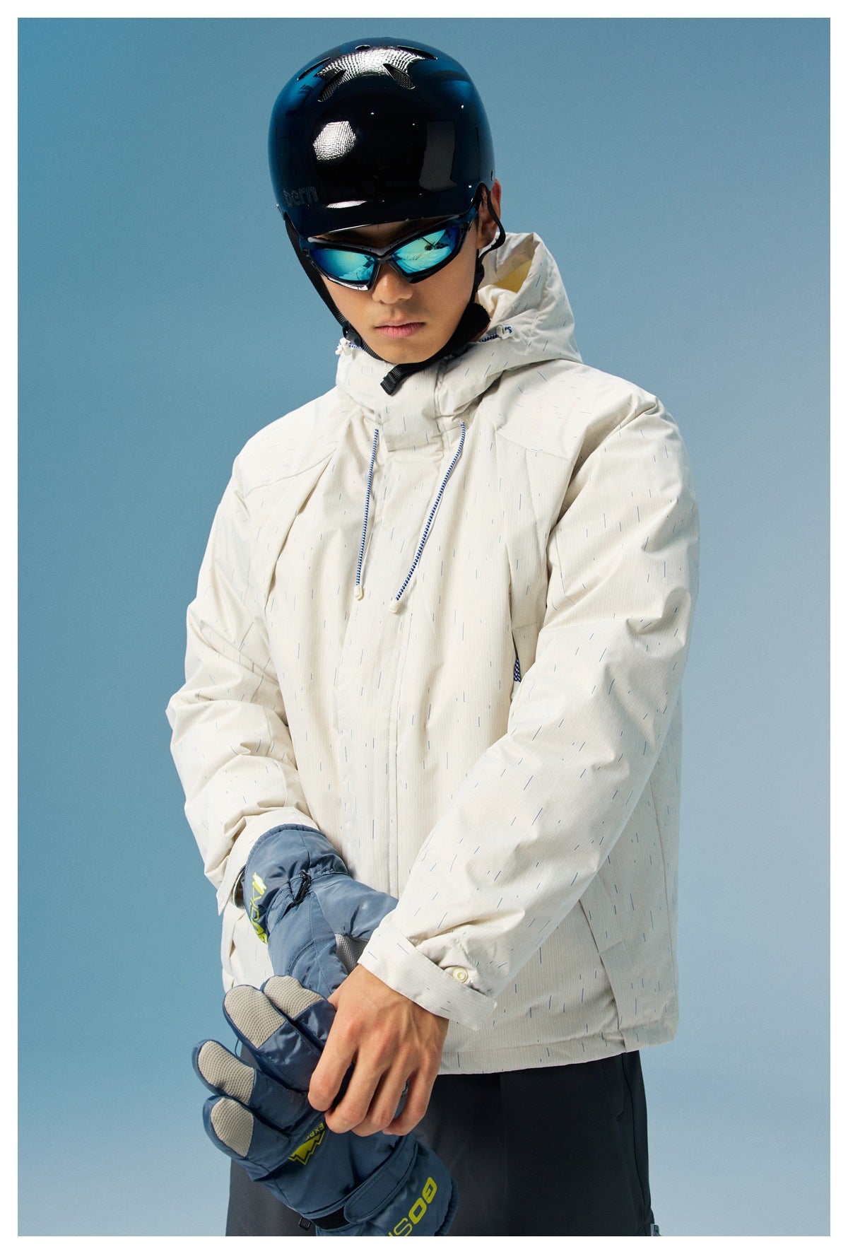 Close view of a model wearing a white patterned hooded ski jacket and helmet, holding blue gloves.