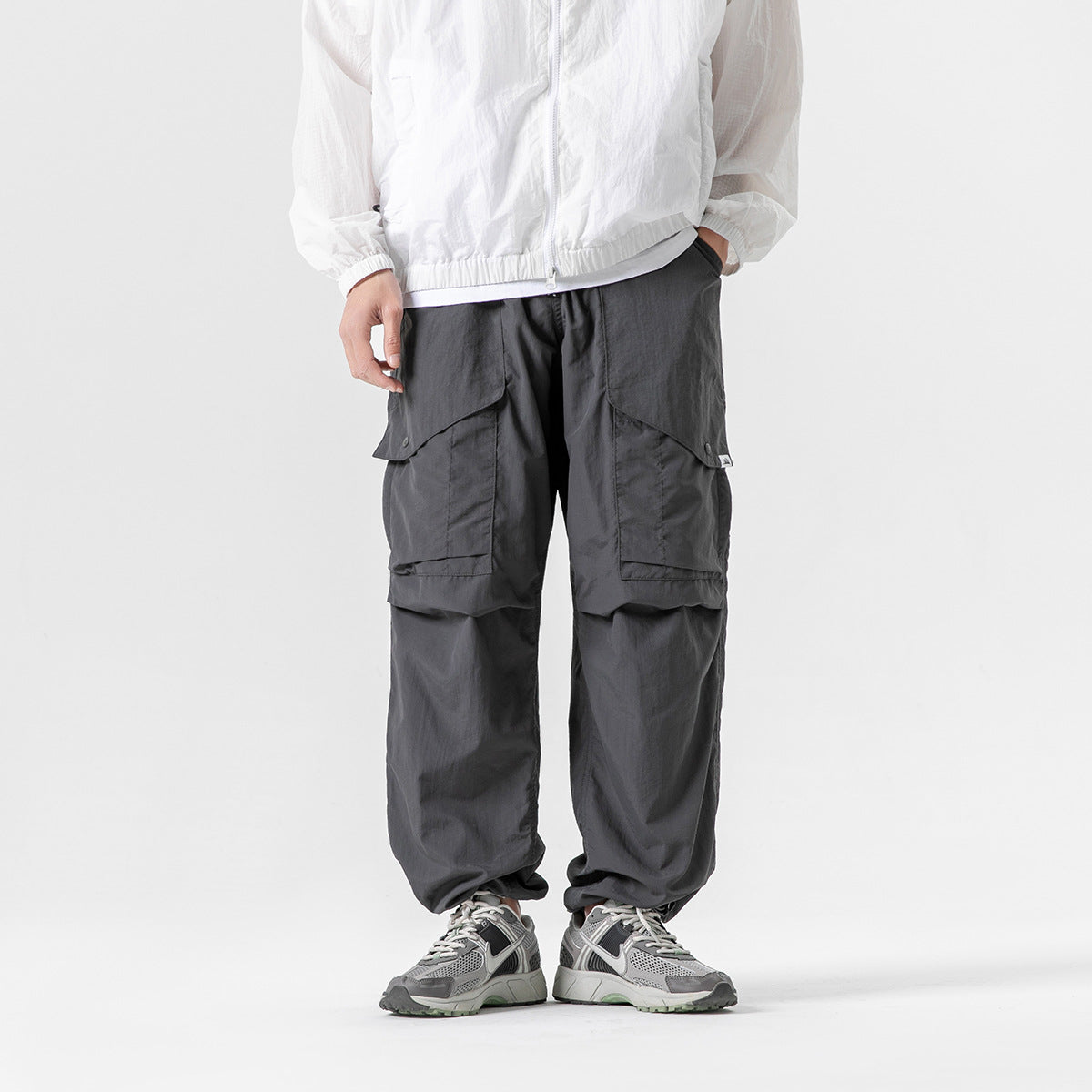 Front view of a model wearing loose charcoal-gray cargo pants with large flap pockets and layered knee panels.