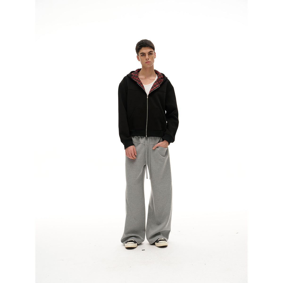 Full-length model wearing a black zip-up hoodie with red plaid hood lining and wide gray sweatpants.