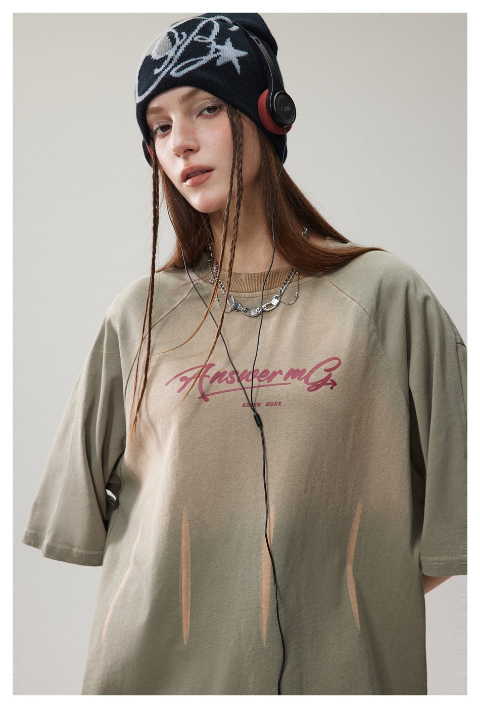 Model wearing olive green faded raglan T-shirt with red script print, beanie, headphones and chain necklace