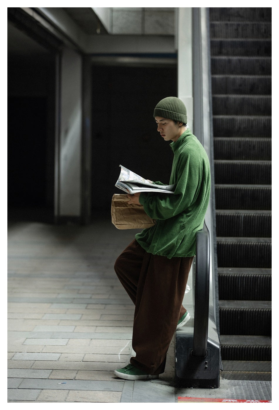 Side view of a person wearing a green quarter-zip pullover and brown wide-leg pants beside an escalator.